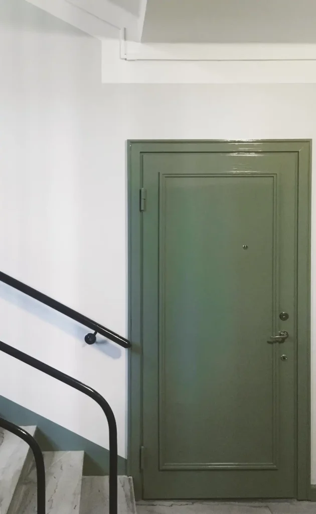 Skogsgrön lägenhetsdörr med dekorativa lister. Dörren har metallhandtag och titthål. Omgiven av ljust målade väggar och svarta räcken vid sidan, med marmor i trappan och på golvet framför.