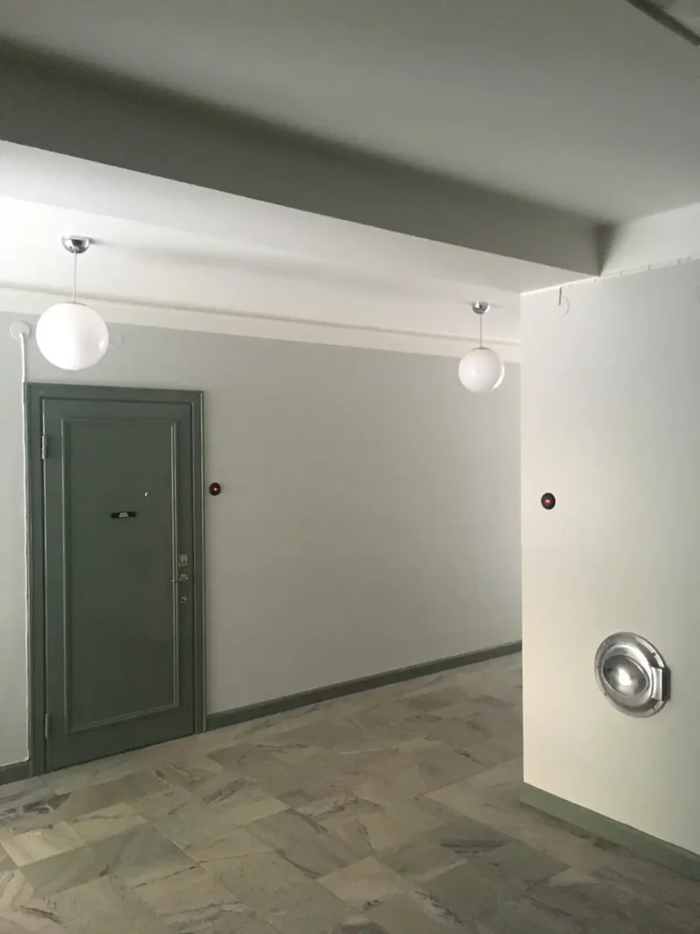 Inre hall med gröngrå lägenhetsdörr till vänster, två vita pendellampor, marmorgolv, runda svarta strömbrytare på väggarna och ett sopnedkast i metall i högra väggen.