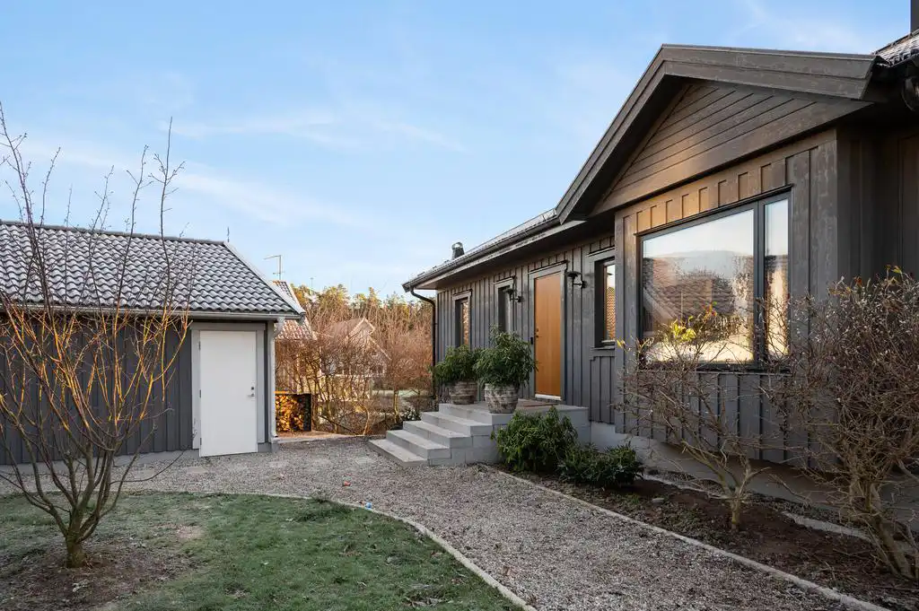 Frontalbild av grått trähus med asymmetriskt gaveltak, träentrédörr och tre trappsteg. Två krukor med växter framför dörren, stort fönster till höger. Intilliggande garage med vit port. Framför huset grusgång och rabatter med buskar.