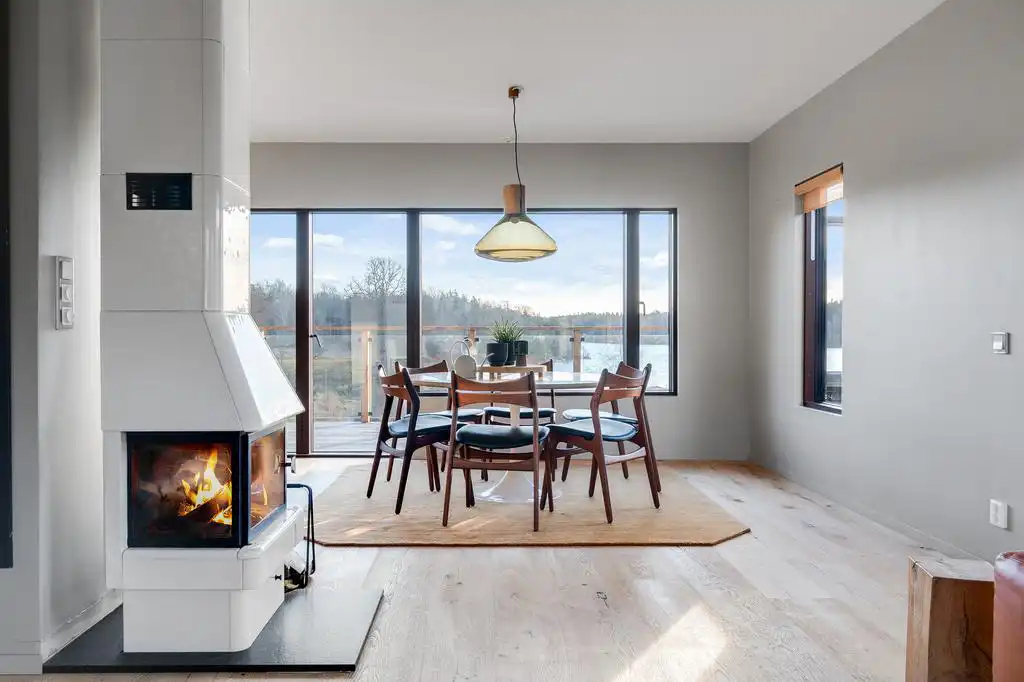 Ljusstark matplats med vita väggar och trägolv. Vit kakelugn med eld i förgrunden. Runt matbord i trä fyra stolar med mörka sitsar. Glaskupolslampa med trädetalj hänger ovanför. Stora fönster visar barrskog och himmel, dagsljus skapar mjuka skuggor. På bordet en kruka med grön växt. Inredningen och eldens sken ger en varm, inbjudande atmosfär.
