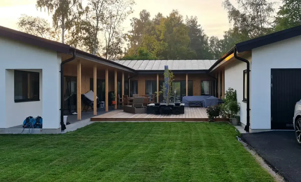 Modern villa med vit puts och träpanel, två enplansflyglar med svart plåttak runt central uteplats med trädäck, loungegrupp och matbord. Till höger garage med svart port och bil. Framför huset gräsmatta, bakom lövträd i sensommarljus.