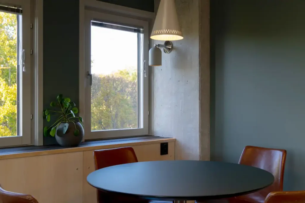 Runt svart mötesbord i förgrunden med tre bruna läderstolar. I bakgrunden två höga fönster mot lövträd, en krukväxt på fönsterbrädet, en betongpelare med vägglampa och en mörkgrön vägg.