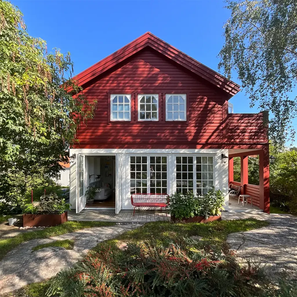Frontalbild av rött tvåvåningsradhus med vitmålade fönster, glasveranda och gaveltak. Framför huset finns grönska, stengång och en röd bänk.