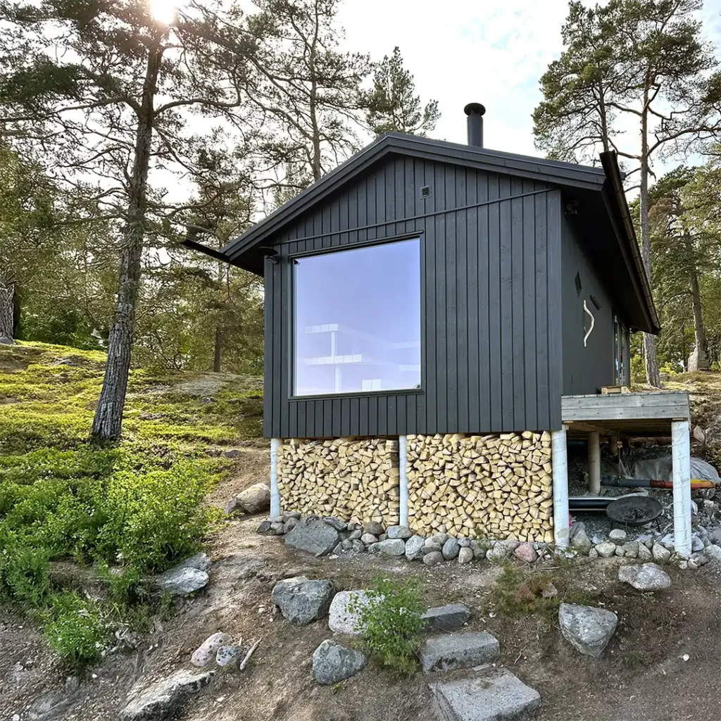 Exteriör vy nerifrån av en bastubyggnad på Värmdö med sadeltak, mörk träpanel och ett stort glasparti. Omkring byggnaden syns en skogsklädd sluttning.