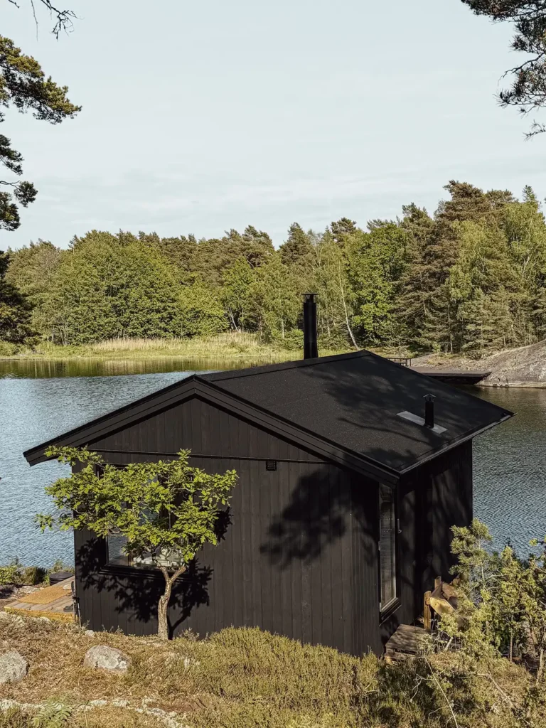 Exteriör vy uppifrån av en bastubyggnad på Värmdö med sadeltak, mörk träpanel och svarta skorstenar. Omkring byggnaden syns en vik, skogsklädda sluttningar och klippor.